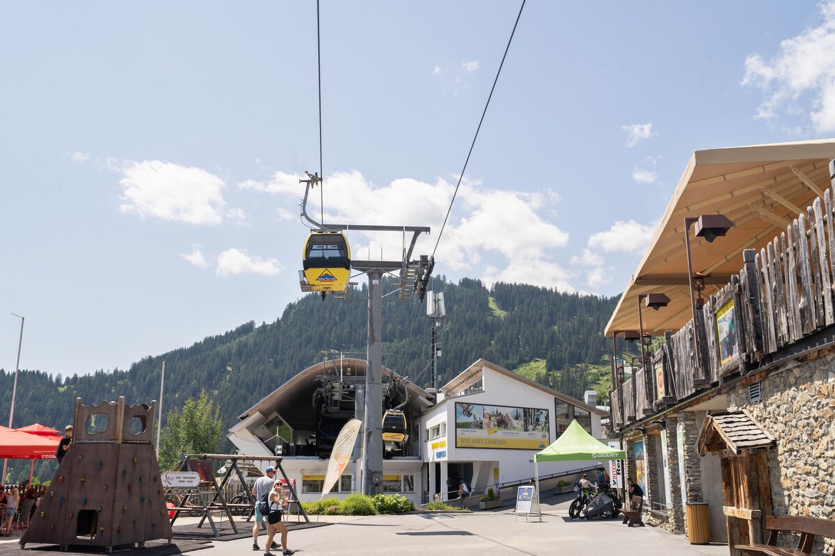 A vibrant outdoor scene is captured, featuring a cable car station against a backdrop of green hills. Near the station, there are wooden structures and colorful canopies providing shade, while leisure activities such as climbing and seating areas enhance the inviting atmosphere.