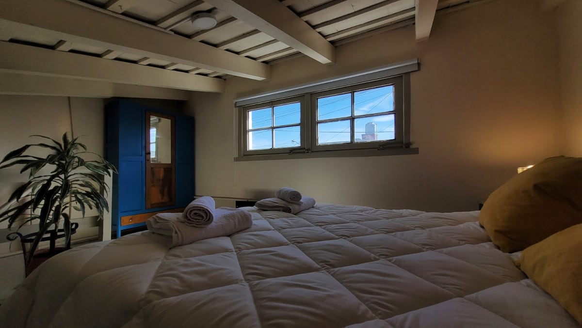 A comfortable sleeping area features a spacious bed covered with a white quilt. Two neatly rolled towels are placed on the bed. Natural light filters through two large windows, highlighting a blue cabinet and a potted plant in the corner.