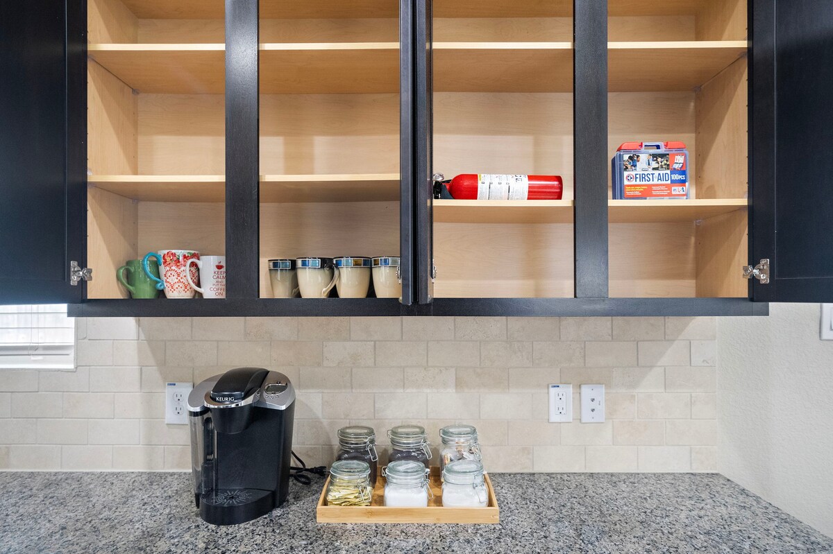 The kitchen features a minimalistic design with open shelving displaying glasses and a first aid kit. A Keurig coffee machine sits next to a wooden tray with glass jars, all set against dark cabinetry and light-colored backsplash.