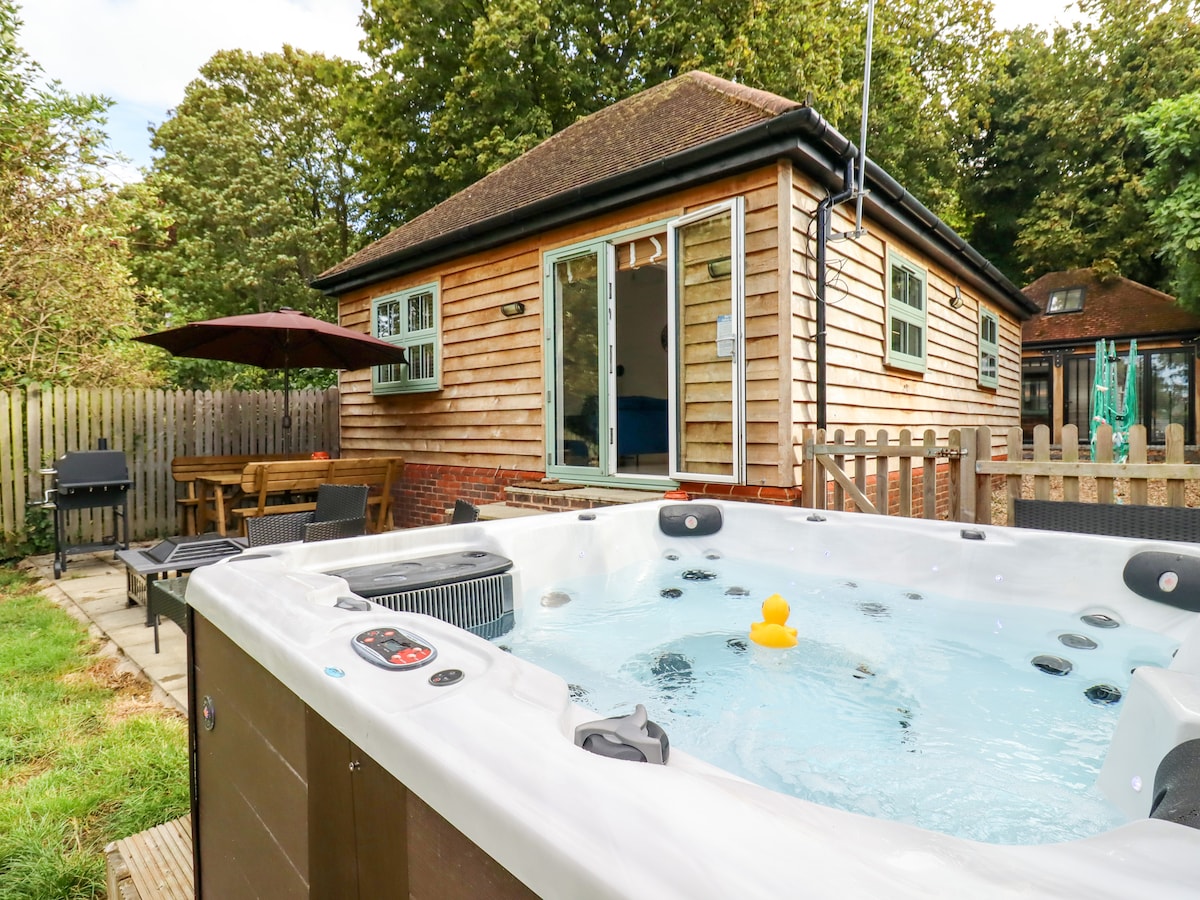 A hot tub with a yellow rubber duck floats on the surface, situated in the foreground. The external annexe is visible in the background, featuring a wooden exterior with large windows. A patio area with outdoor furniture and a barbecue is surrounded by greenery.