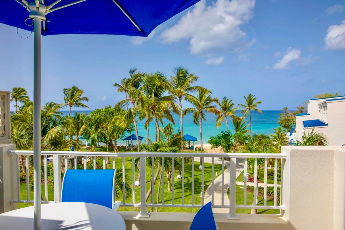 A balcony view showcases vibrant blue umbrellas and lush palm trees overlooking a serene ocean. The exterior table and chairs are positioned for relaxation, complementing the tranquil scenery of the beach and tropical landscape.