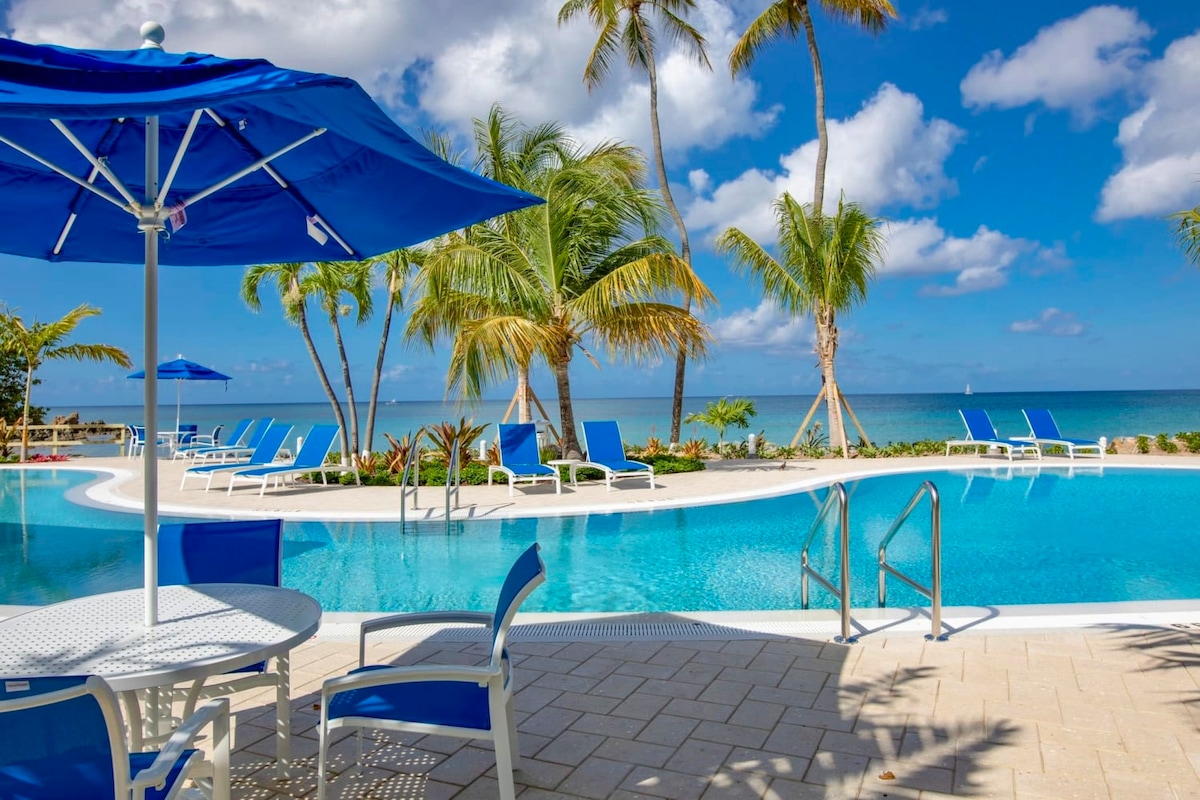 An outdoor swimming pool is surrounded by palm trees and lounge chairs. Blue umbrellas provide shade over white patio furniture. The clear water reflects the blue sky and nearby ocean, creating a serene atmosphere for relaxation.