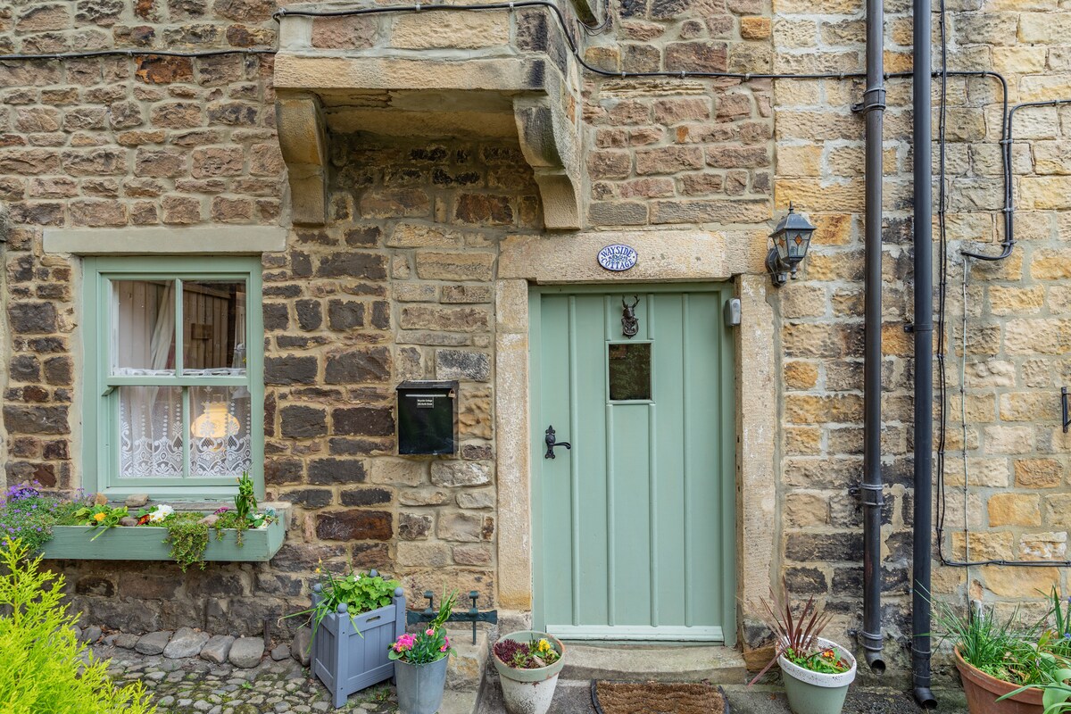 The cottage exterior features a charming stone facade with a light green door at the center. Flower pots adorn the entrance, and a traditional lantern adds to the quaint atmosphere. A small window with lace curtains is visible to the left.