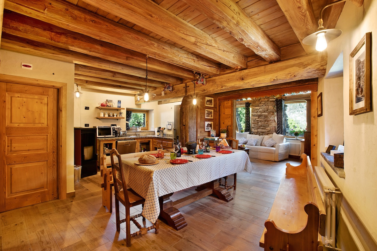 An open-plan living space features exposed wooden beams and a large dining table set with tableware. A fully-equipped kitchen is visible to the left, while a cozy seating area with a sofa is in the background, framed by windows showcasing the garden view.