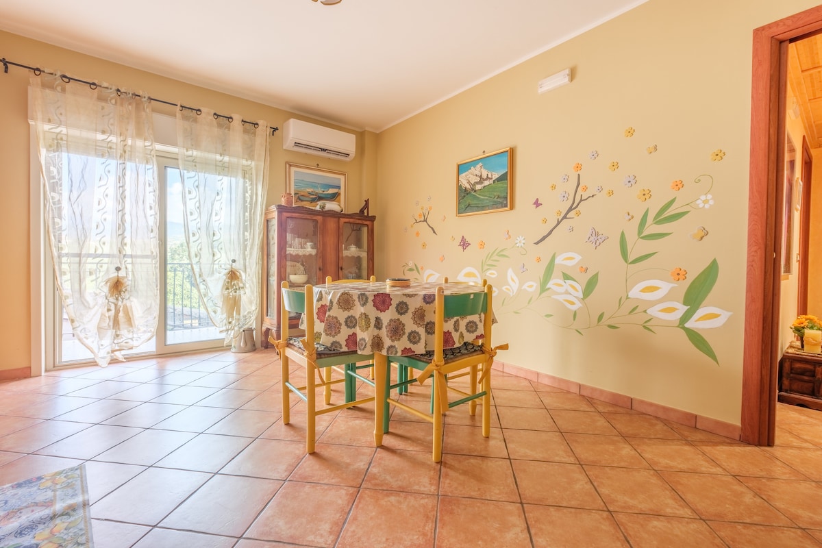 A dining area is presented with a colorful table surrounded by chairs. Large windows allow natural light to illuminate the space, and a mural of flowers adds a touch of decor. Air conditioning is mounted on the wall, and a cabinet is visible in the corner.
