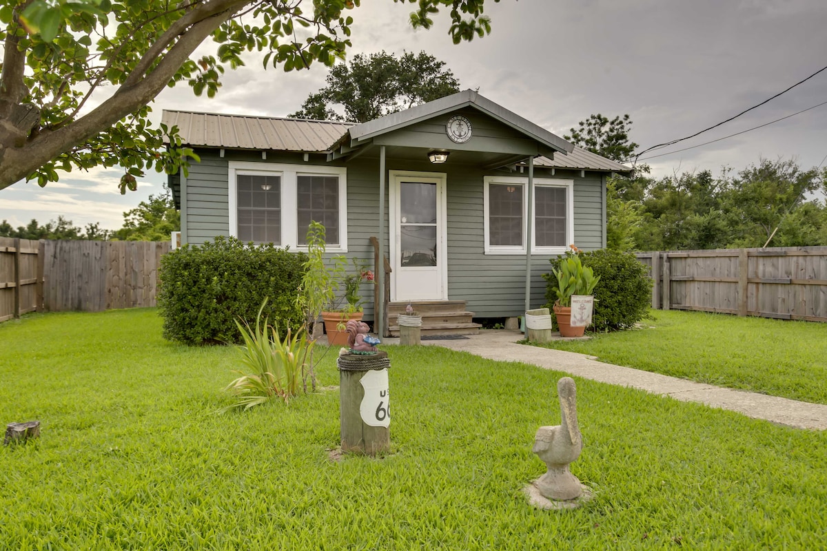 The exterior of a charming gray cottage is framed by a well-manicured green lawn. A wooden pathway leads to the entrance, bordered by potted plants and a decorative stone. A clock adorns the facade, contributing to the inviting atmosphere. The property is enclosed by a wooden fence.
