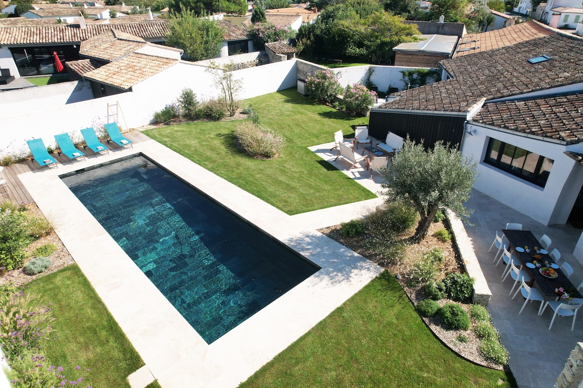 An aerial view of a spacious garden features a heated outdoor swimming pool bordered by a stone terrace. Sun loungers are arranged along one side of the pool, while a dining area with seating is positioned on the other, surrounded by well-kept greenery.