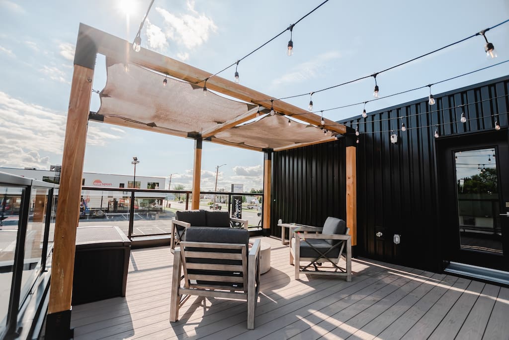 A rooftop patio features wooden beams and a canopy, providing shade over outdoor seating arrangements. Two lounge chairs and a circular table are positioned for relaxation. String lights hang overhead, adding subtle illumination during evening hours.