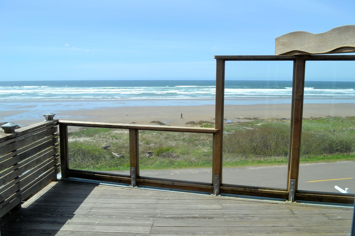A glassed deck offers unobstructed views of the ocean and sandy beach below. The horizon is lined with rolling waves under a clear blue sky, while a person is seen walking along the shore. The deck's wooden railing complements the natural setting.