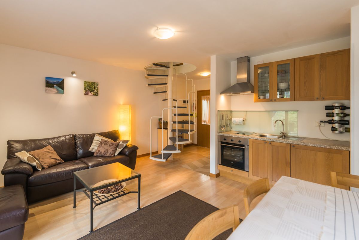 An open plan living area features a dark leather sofa and a coffee table, complemented by a dining table with wooden chairs. A spiral staircase leads to the upper level. The kitchen area includes modern appliances and cabinetry, illuminated by natural light from nearby windows.