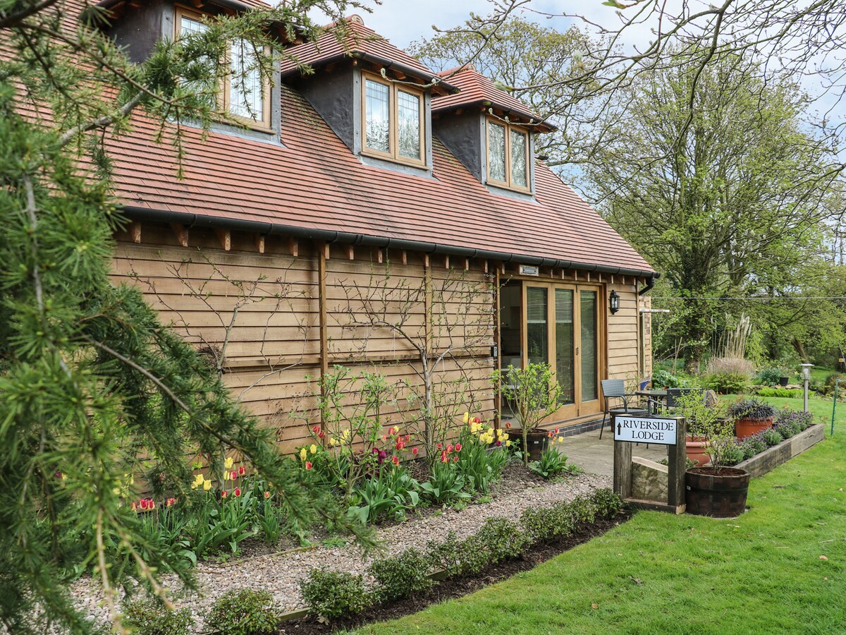 The exterior of a charming wooden cottage is visible, featuring a sloped roof and large glass doors. A well-maintained garden with vibrant tulips and greenery surrounds the entrance, while a sign marked 'Riverside Lodge' is prominently displayed at the front.