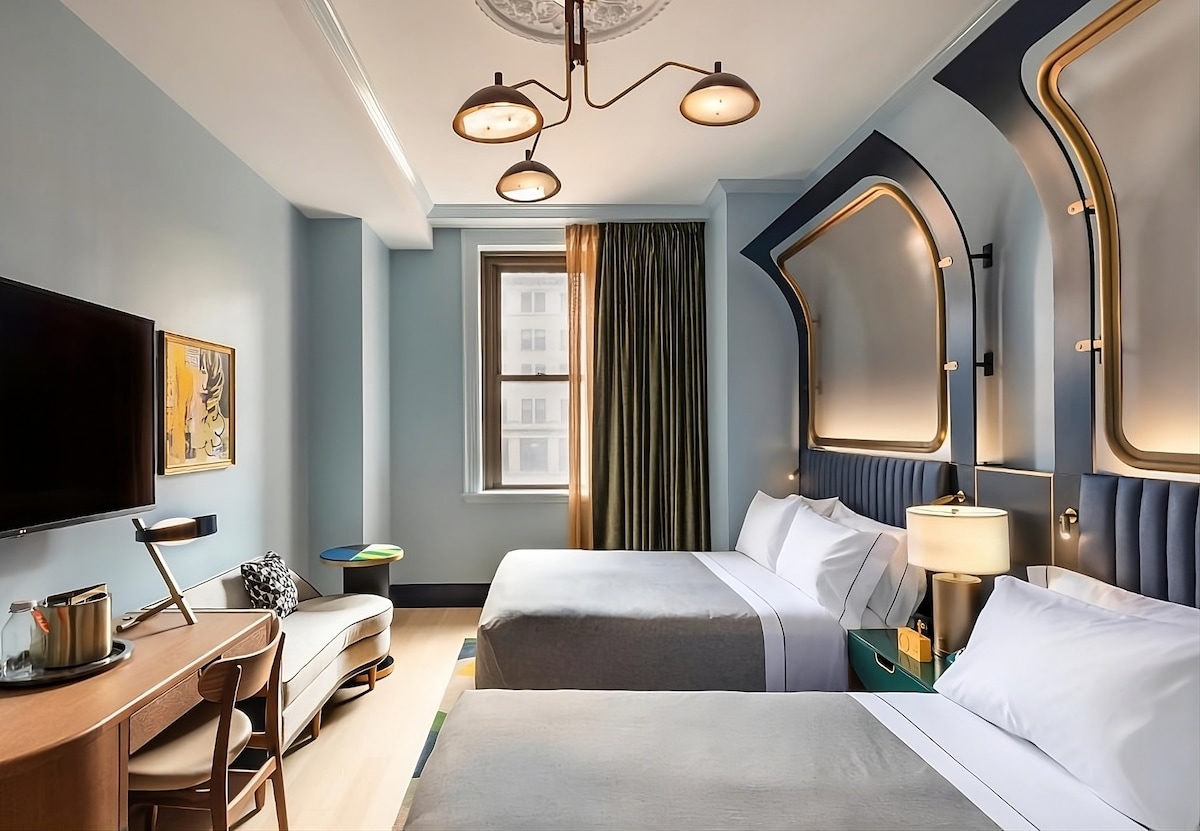 A well-appointed hotel room showcases two spacious queen beds with soft bedding. A flat-screen TV is mounted on the wall opposite a wooden work desk and chair. The room is bathed in natural light from a large window adorned with green curtains, complemented by stylish lighting fixtures.