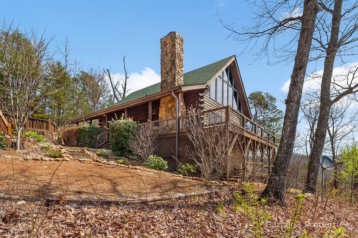 The exterior of a spacious log cabin is visible, featuring a prominent stone chimney and a green roof. Surrounding foliage includes trees and shrubs, with a wide deck accessed by steps. Natural ground cover consists of leaf litter and stones, enhancing the mountain retreat ambiance.