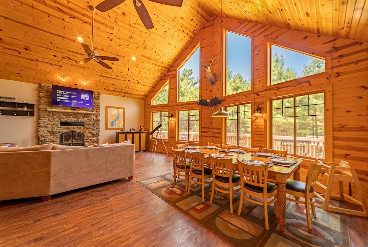 A spacious living area with vaulted wooden ceilings features large windows that offer abundant natural light. A stone fireplace serves as a focal point, while a dining table with multiple chairs invites gatherings. The warm wood paneling enhances the cabin's rustic charm.