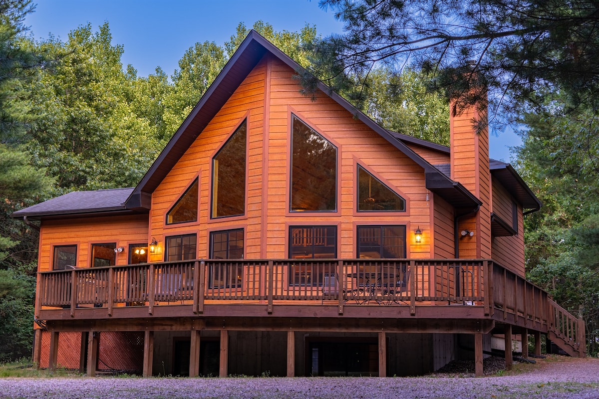 The cabin's exterior showcases a striking combination of wood and large triangular windows under a clear blue sky. A spacious deck wraps around the structure, providing access to the serene forest surroundings. Pine trees frame the scene, emphasizing the cabin's natural setting.