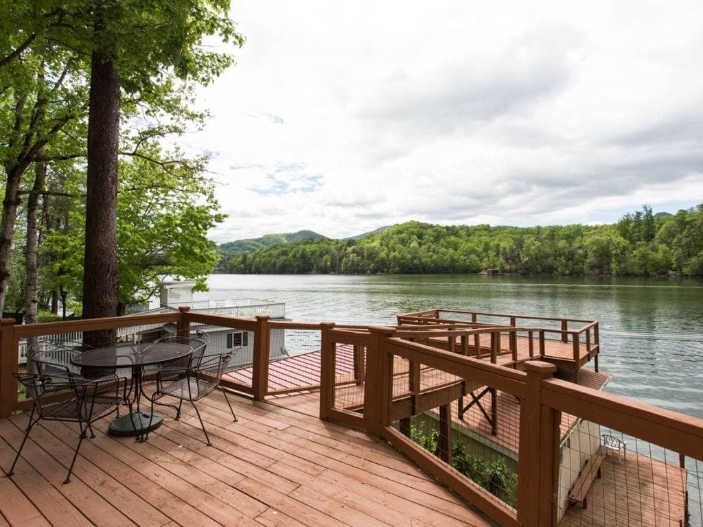 A spacious deck is seen overlooking the calm waters of the lake, framed by lush green trees. A table with four chairs is positioned for gatherings, and the surrounding scenery showcases the gentle slopes of the mountains in the distance under a partly cloudy sky.