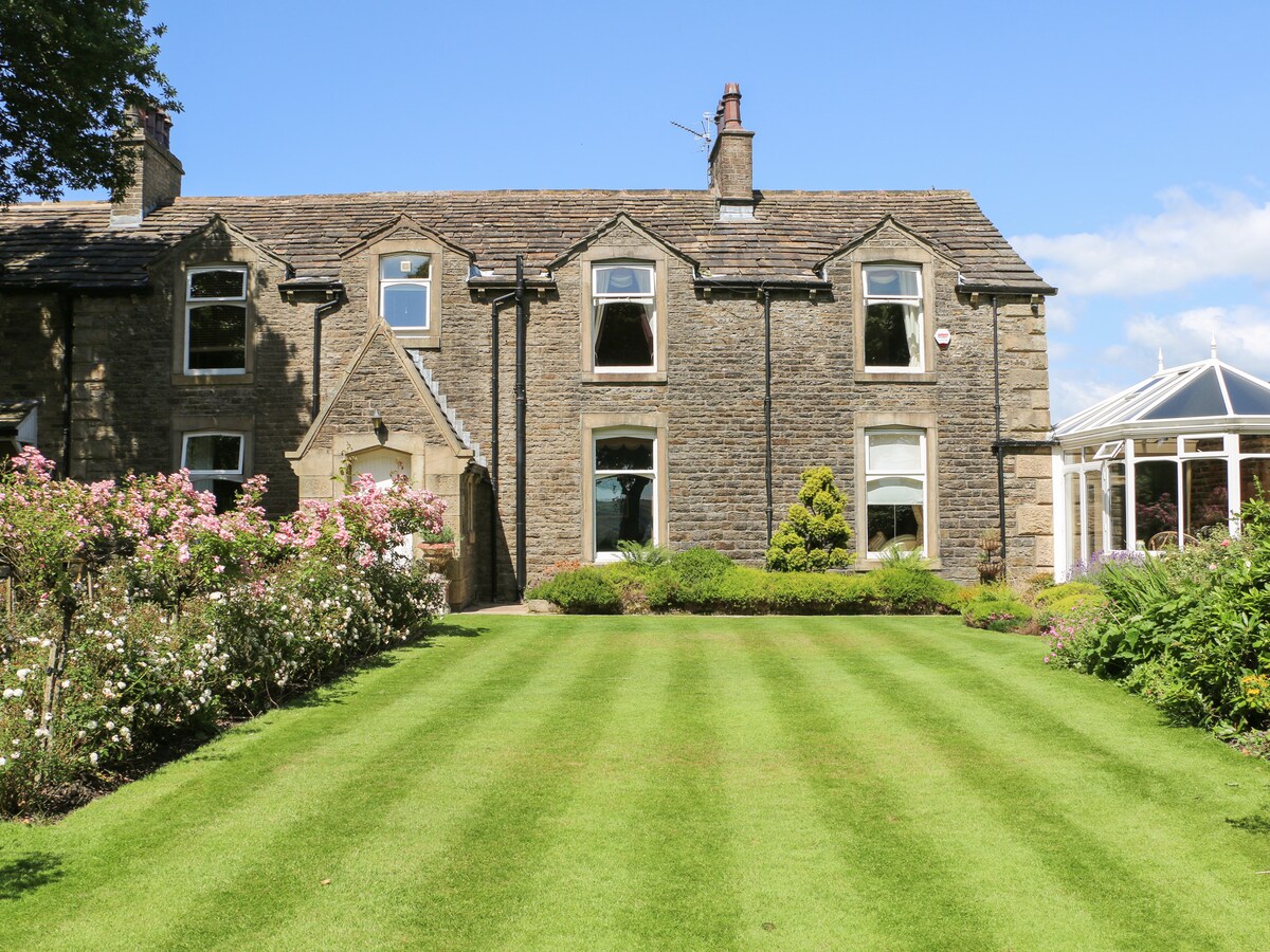 The exterior of the cottage features a well-maintained garden with striped green lawns and blooming rose bushes. Large windows reflect natural light, showing the inviting architecture of the stone building. A conservatory can be seen on the right, providing a glimpse of additional living space.