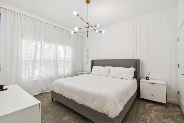 Este dormitorio luminoso y elegante te invita a relajarte con su lujosa cama tapizada gris y su suave alfombra. Un candelabro contemporáneo añade estilo, mientras que las cortinas blancas transparentes enmarcan la ventana, creando un ambiente aireado.