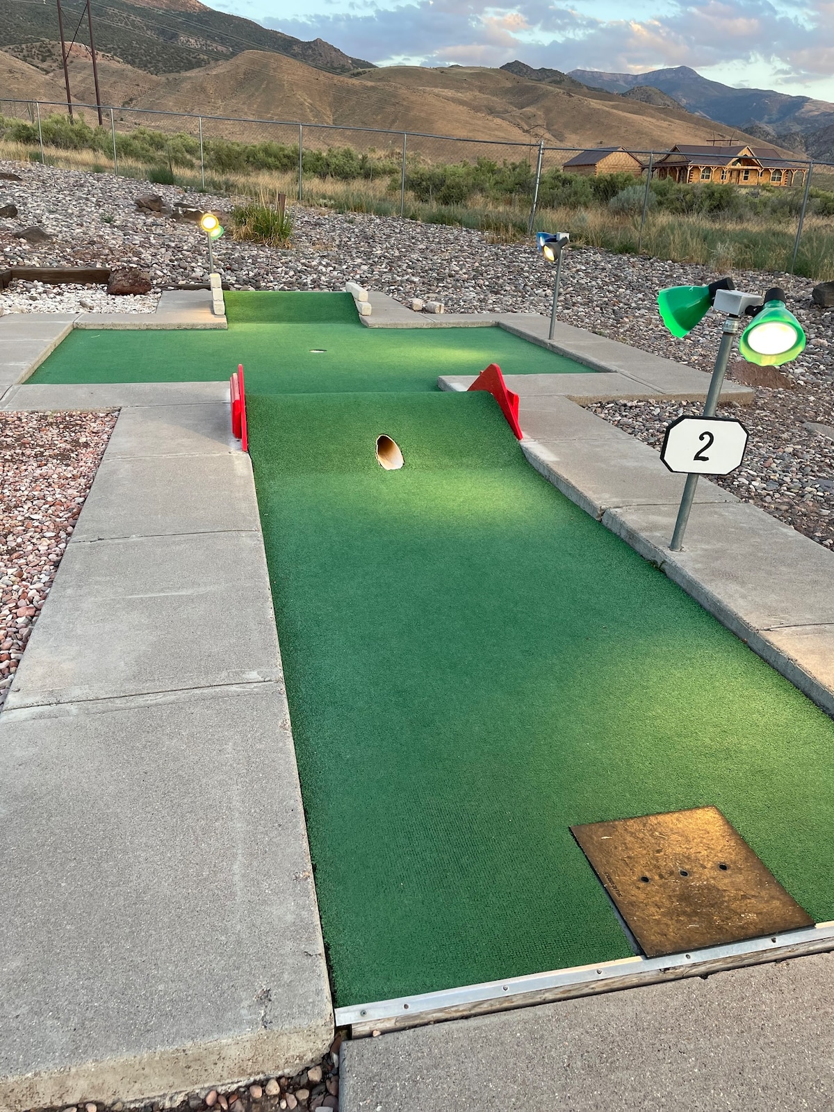 A miniature golf course is presented with two putting lanes marked by numbered signs. The vibrant green carpet covers the ground, while strategically placed color-lit lamps provide illumination. Rocky terrain and a mountainous landscape form the backdrop, enhancing the outdoor setting.