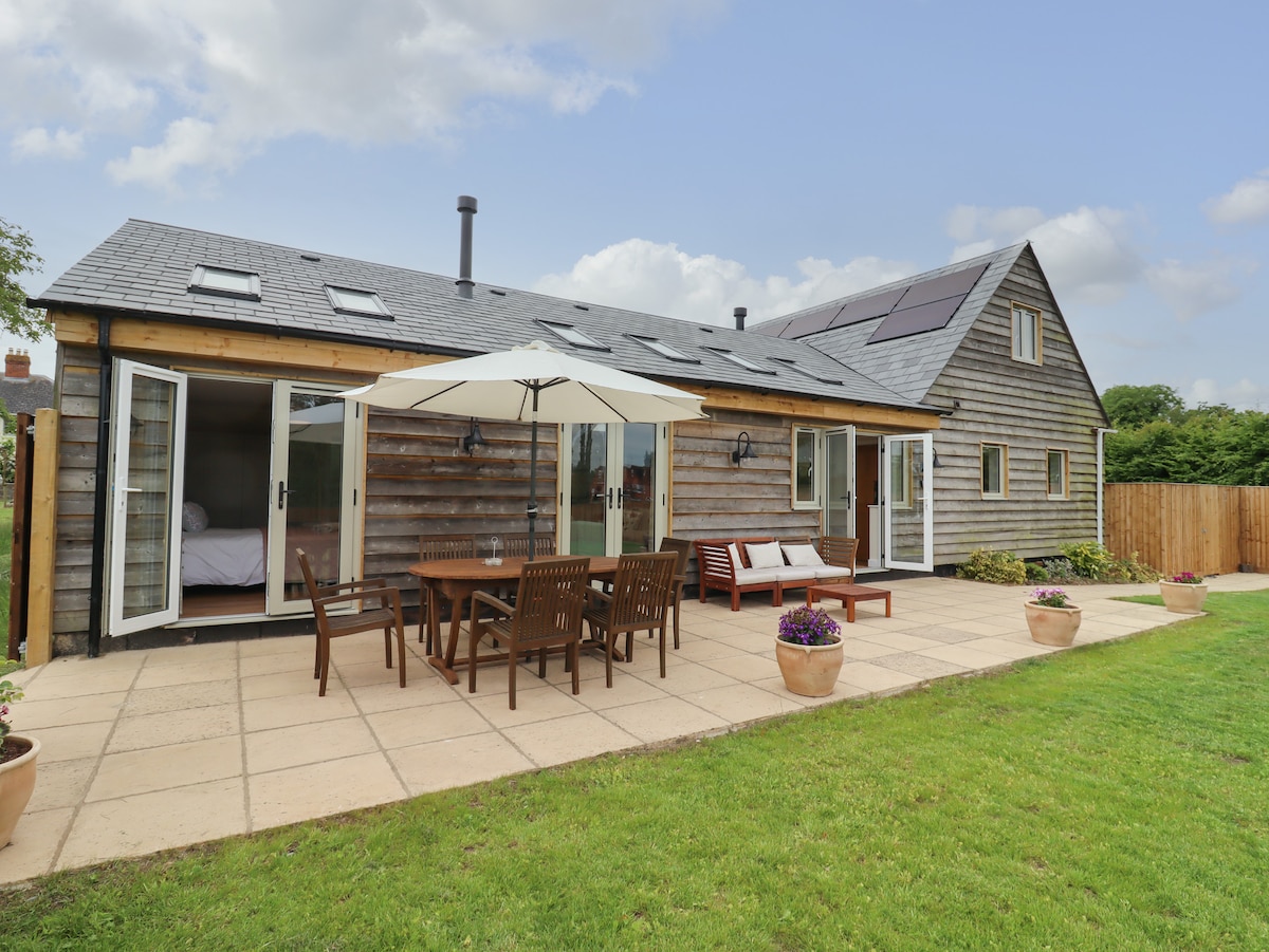 The exterior of the bungalow displays a wooden facade and a spacious patio area. A large dining table with matching chairs is positioned under a parasol, accompanied by potted plants. Large glass doors open to reveal the interior, with a well-maintained lawn beyond.