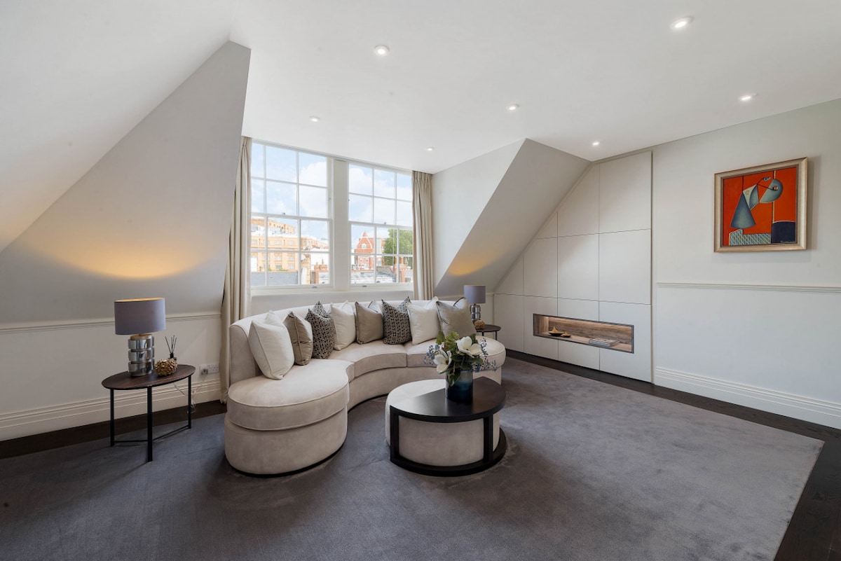 A spacious lounge area features a comfortable curved sofa adorned with decorative pillows. Natural light fills the space through large windows, while soft lighting is provided by two lamps. An elegant wall art piece adds a touch of color above a modern fireplace, complementing the neutral decor.