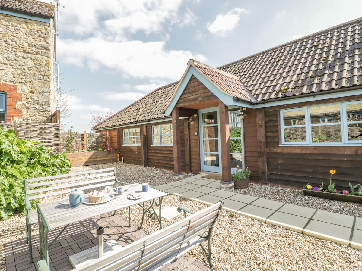 The exterior of the barn features a charming wooden structure with a slate roof. A spacious patio area with a table and seating is visible, surrounded by gravel and potted plants. Large windows allow natural light to illuminate the entrance.