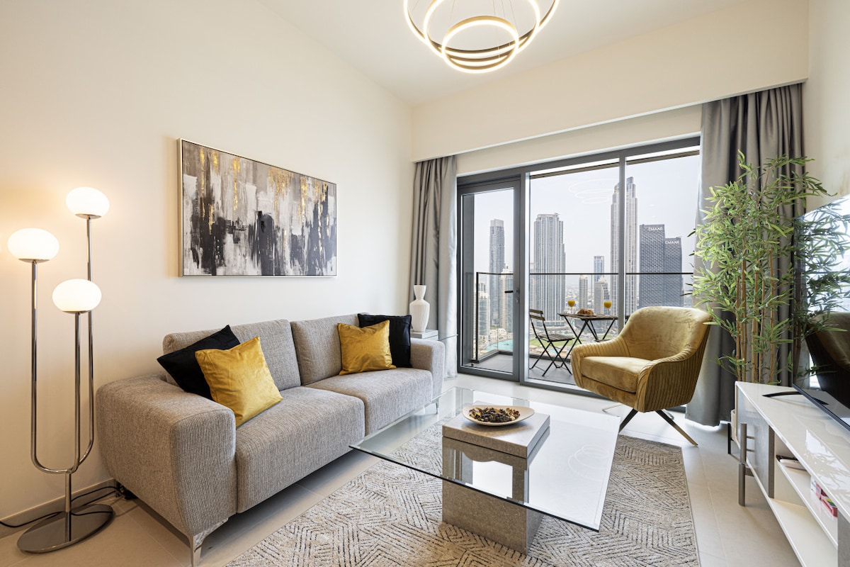 A modern living space features a plush gray sofa adorned with yellow accent cushions, complemented by a glass coffee table. A yellow armchair adds a pop of color. Expansive windows frame a city skyline view, while a stylish lamp and decorative plant enhance the ambiance.