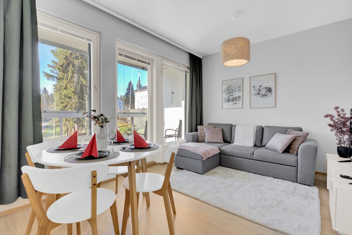 The living area features a grey sectional sofa paired with a soft rug. A round dining table is set for four, with vibrant red napkins and a vase of flowers at the center. Large windows invite natural light, providing a view of the outdoors.