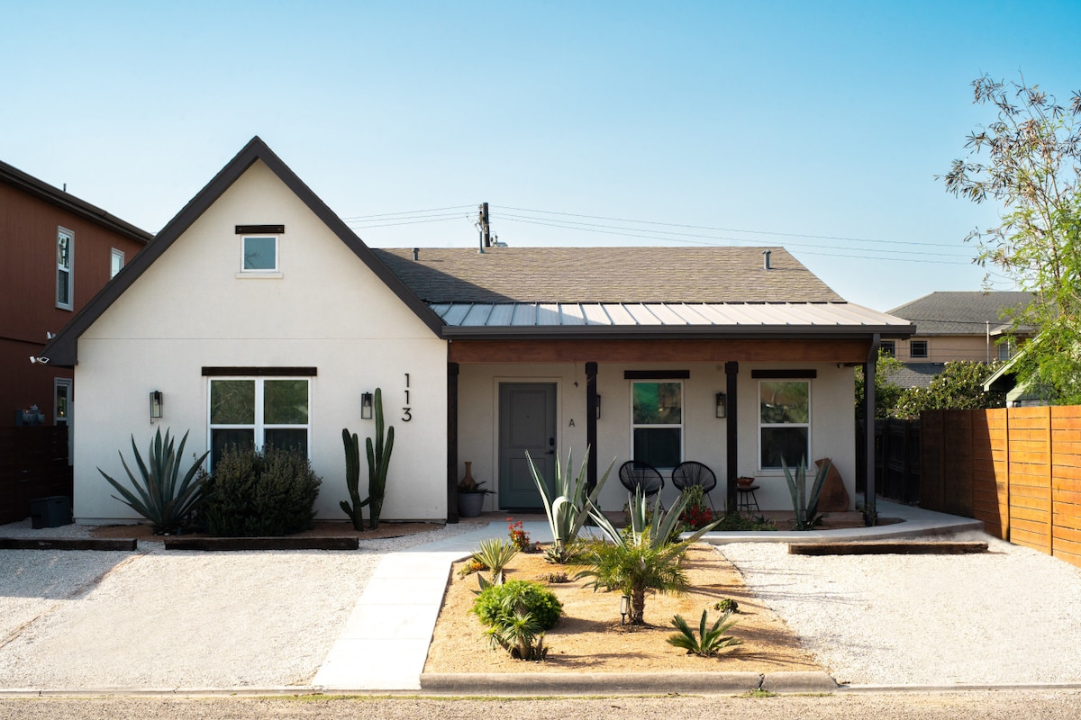 A modern duplex exterior featuring a white facade with dark trim and a welcoming front porch. The landscape includes sandy gravel and drought-resistant plants, such as tall cacti and palm trees. Two black chairs are positioned near the entrance, adding an inviting touch to the outdoor space.