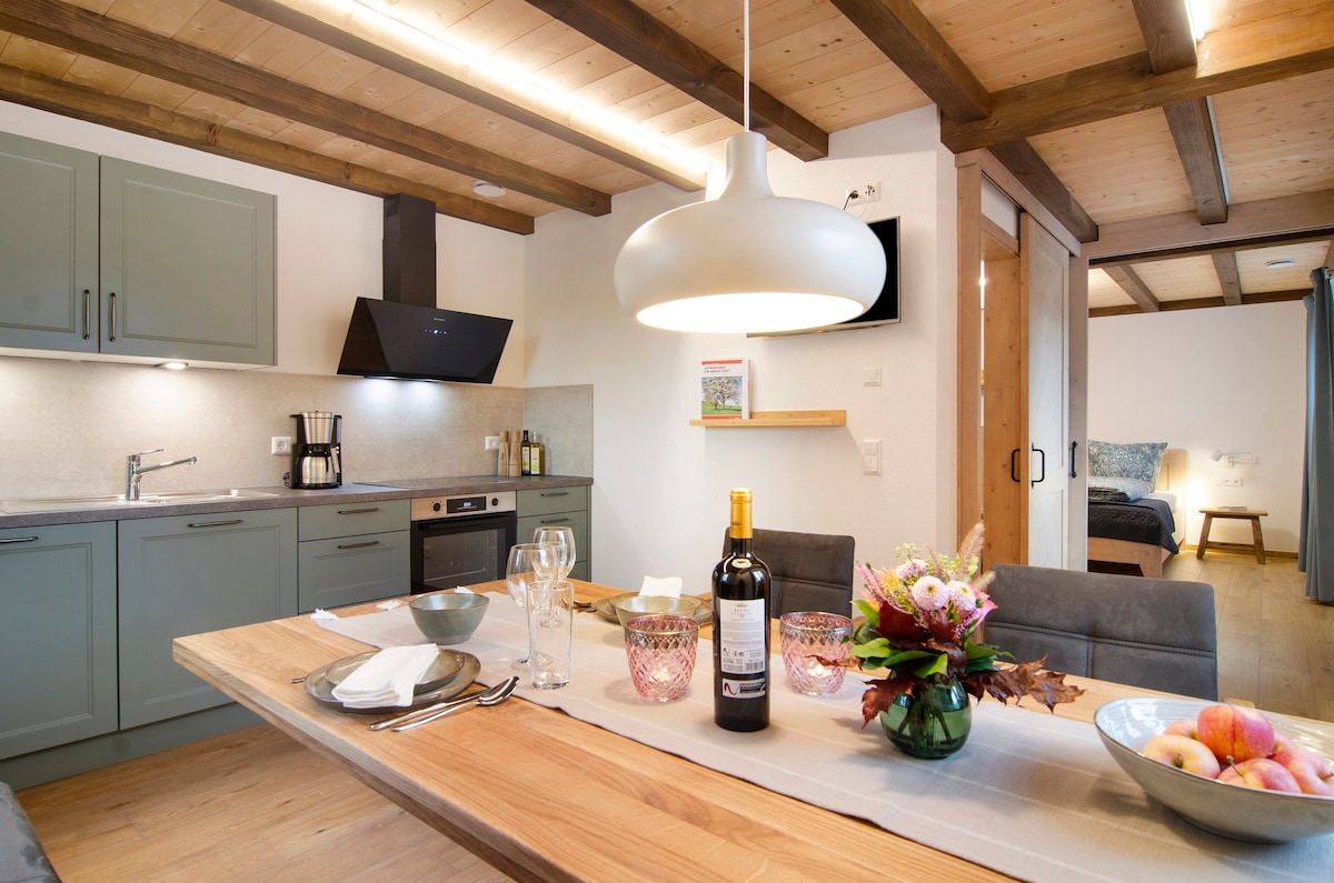 A stylish kitchen and dining area feature modern gray cabinetry and a wooden dining table set for a meal. A bottle of wine and glasses are placed on the table, complemented by fresh flowers and fruit. Soft lighting enhances the warm wooden beams and cozy atmosphere.