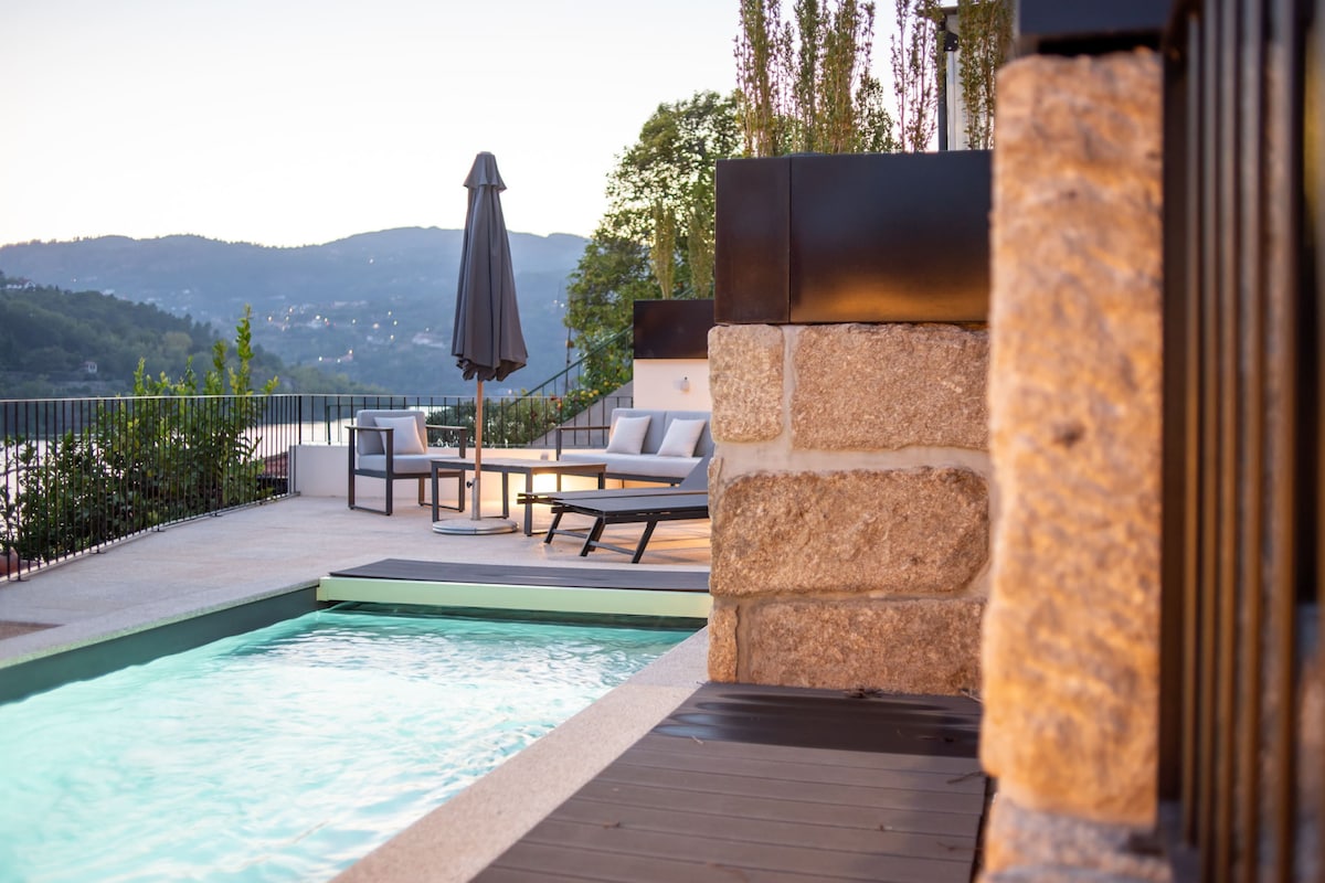 A small heated pool is framed by a stone wall and a wooden deck, offering a serene view of the surrounding hills. A gray umbrella provides shade over a lounge area equipped with modern outdoor furniture, inviting relaxation by the water under the open sky.