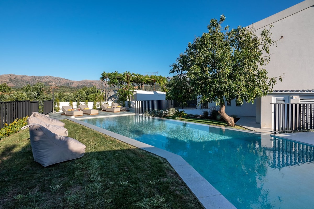 A private pool features a clear blue surface, surrounded by landscaped green grass. Comfortable outdoor seating areas are arranged nearby, inviting relaxation. Lush plants and a tree provide shade, while a view of distant mountains enhances the tranquil setting.