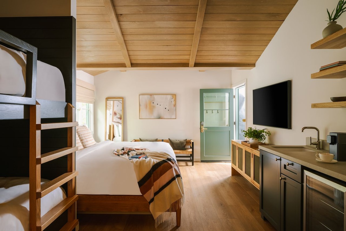 A cozy room features a spacious bed and a set of wooden bunk beds. Natural light enters through large windows, illuminating the light-colored walls and wood ceiling. A small kitchenette is seen along one wall, complemented by a flat-screen TV and stylish shelving.