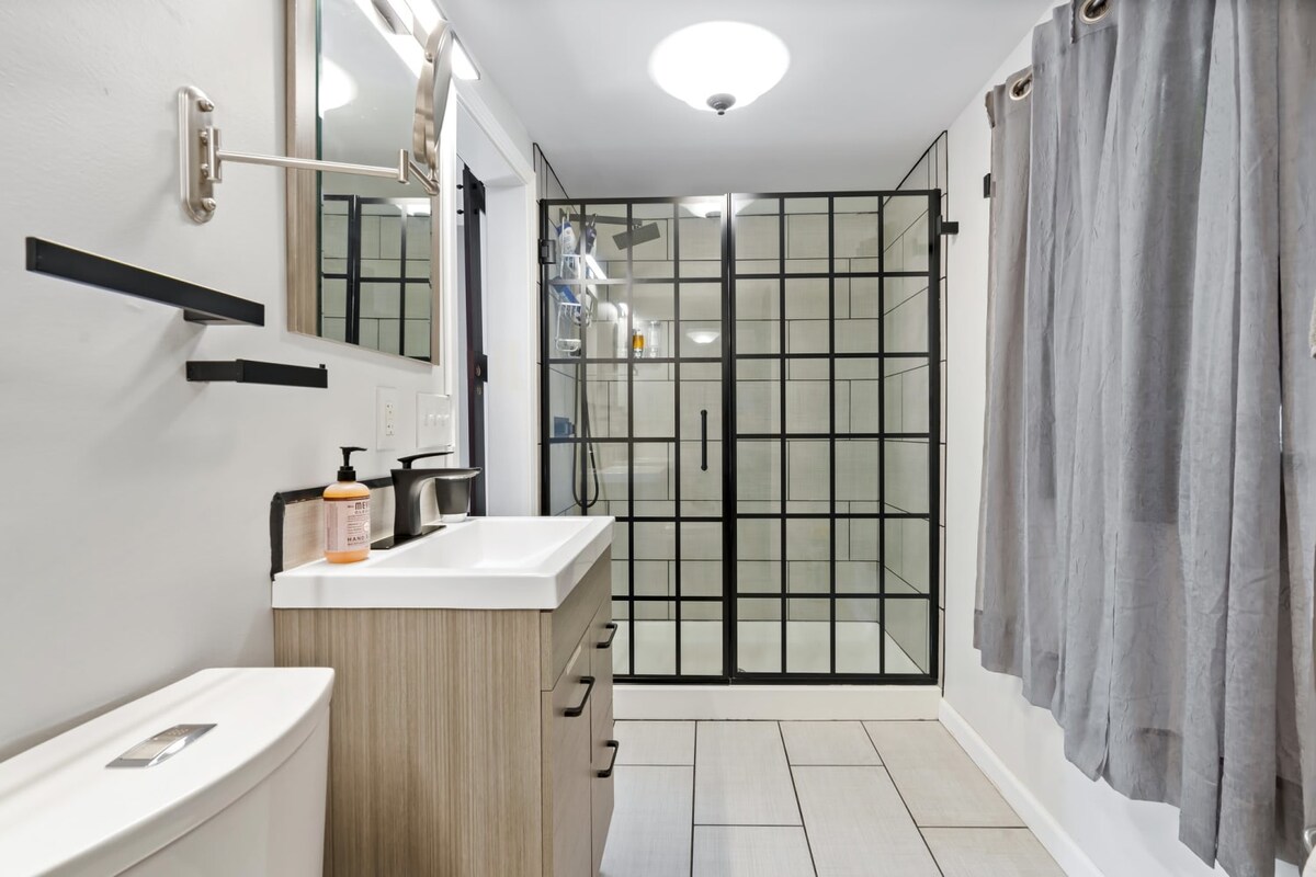 A modern bathroom features a sleek, white vanity and a large glass shower enclosed in black metal framing. Neutral tiles create a clean aesthetic, while a dark curtain adds a touch of privacy. A round ceiling light brightens the space.
