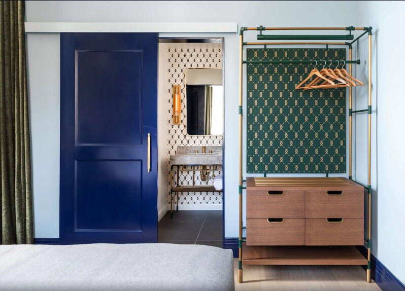 An open hotel room is showcased, featuring a modern wardrobe with hangers and a wooden drawer unit. A stylish blue door leads to a bathroom, reflected in a neutral color scheme. The area is complemented by patterned wallpaper, enhancing the overall aesthetic.