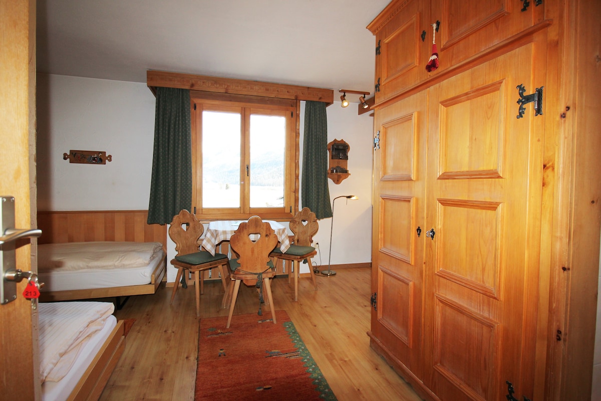 A spacious studio interior features light wooden decor, including a large wardrobe and a dining area with wooden chairs. Two single beds are positioned near a window, with green curtains providing a touch of color. A warm, inviting rug lies on the wooden floor.
