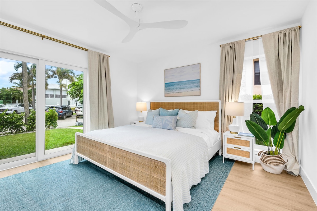 A bright bedroom features a spacious bed with a woven cane headboard, accompanied by soft blue decorative pillows. Light floods through a sliding glass door, which opens to a view of landscaped greenery. White curtains frame the window, enhancing the airy ambiance.