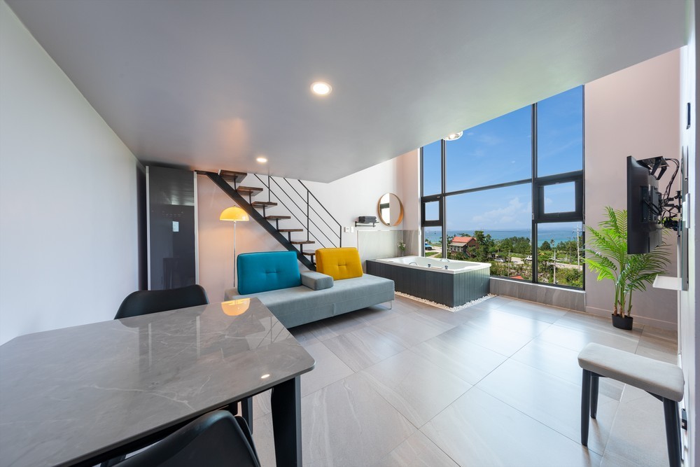 A modern living area features a staircase leading to an upper level. A gray sofa and a blue accent chair complement a bright space illuminated by large windows. A small dining table is positioned nearby, with a spa visible in the background.