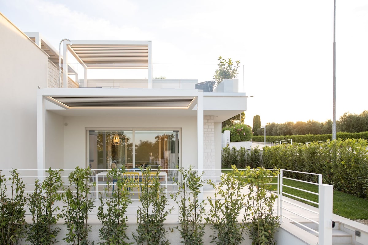 The exterior view of the villa showcases a modern design with large glass sliding doors leading to a terrace. Lush greenery borders the property, while a spacious outdoor seating area is visible, creating a seamless connection between indoor and outdoor living.