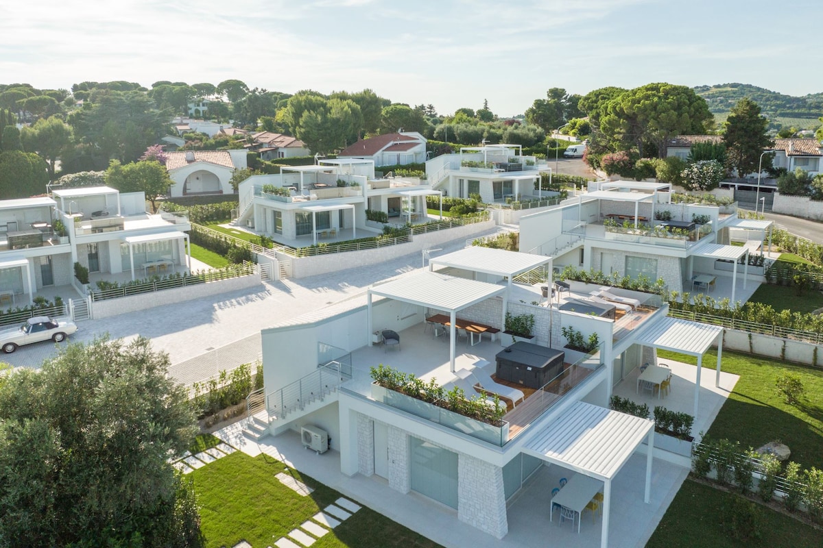 Aerial view of a modern villa complex featuring multiple semi-detached villas set within landscaped gardens. Each villa boasts outdoor terraces, and the paths are lined with greenery, offering a serene and spacious environment. Ample parking spaces are visible, contributing to the layout.