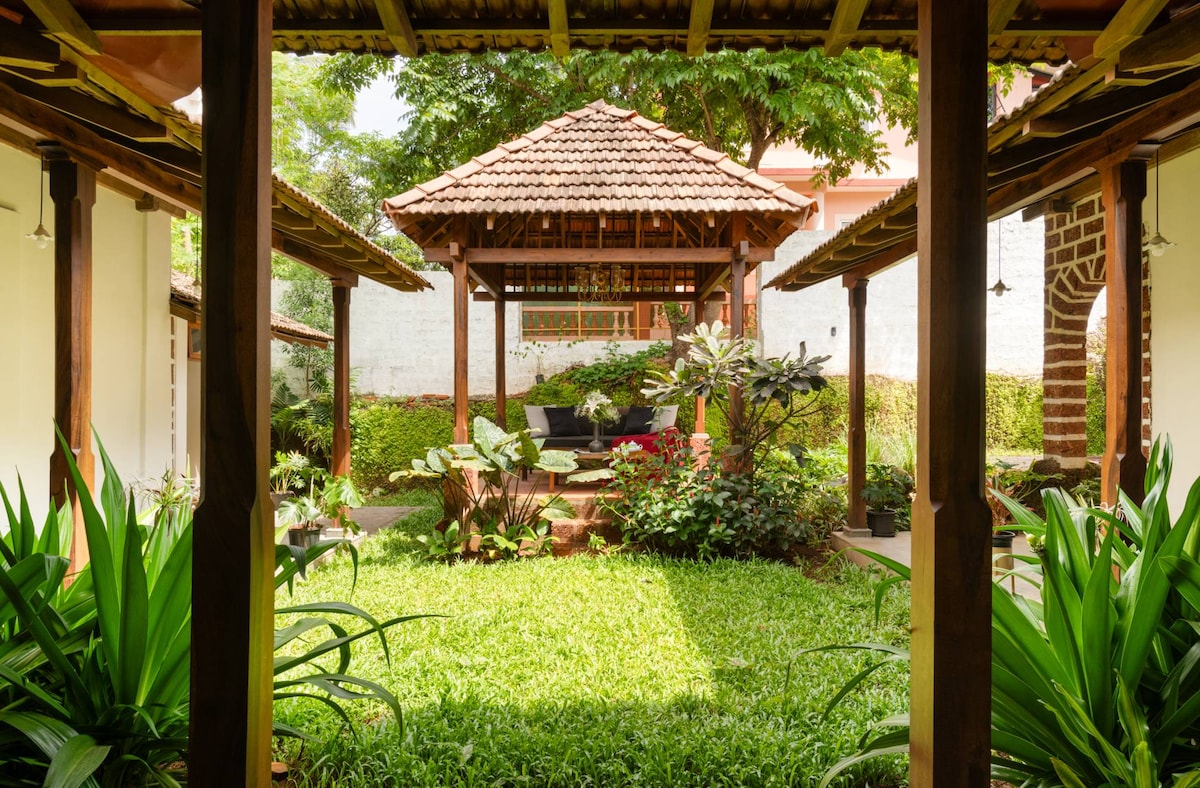 A peaceful courtyard is framed by traditional architecture, showcasing a gazebo with comfortable seating. Lush greenery, vibrant plants, and flowers surround the area, creating a tranquil environment. Natural light filters in, enhancing the calm ambiance of this garden retreat.