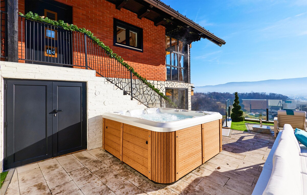 An outdoor area features a wooden hot tub beside a well-maintained stone and tile patio. A staircase leads to an upper level, framed by black railings and adorned with greenery. Clear skies and a scenic view of the landscape are visible in the background.