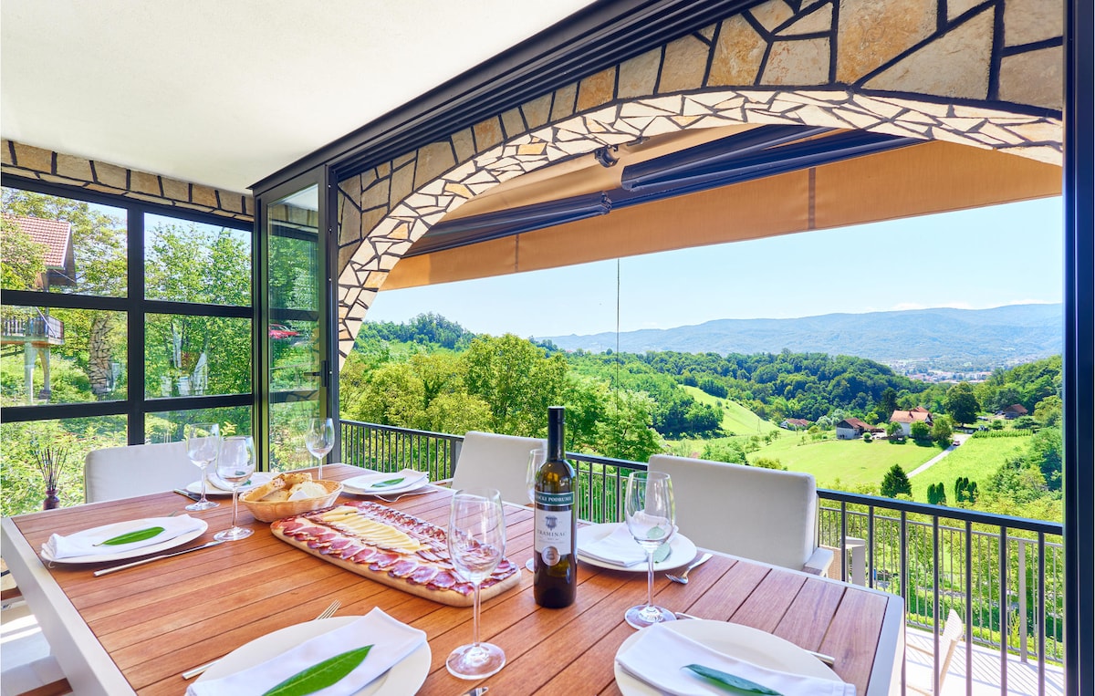 An outdoor dining area features a wooden table set with plates and glasses, overlooking lush green hills. A bottle of wine is placed at the center, while the arched awning provides shade. Sliding glass doors frame the picturesque view of the surrounding landscape.