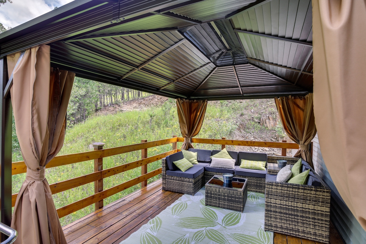 A shaded outdoor pavilion features comfortable wicker seating adorned with decorative cushions. The wooden deck stretches into a serene green landscape, with gentle hills visible in the background. Natural light filters through draped curtains, enhancing the cozy atmosphere.