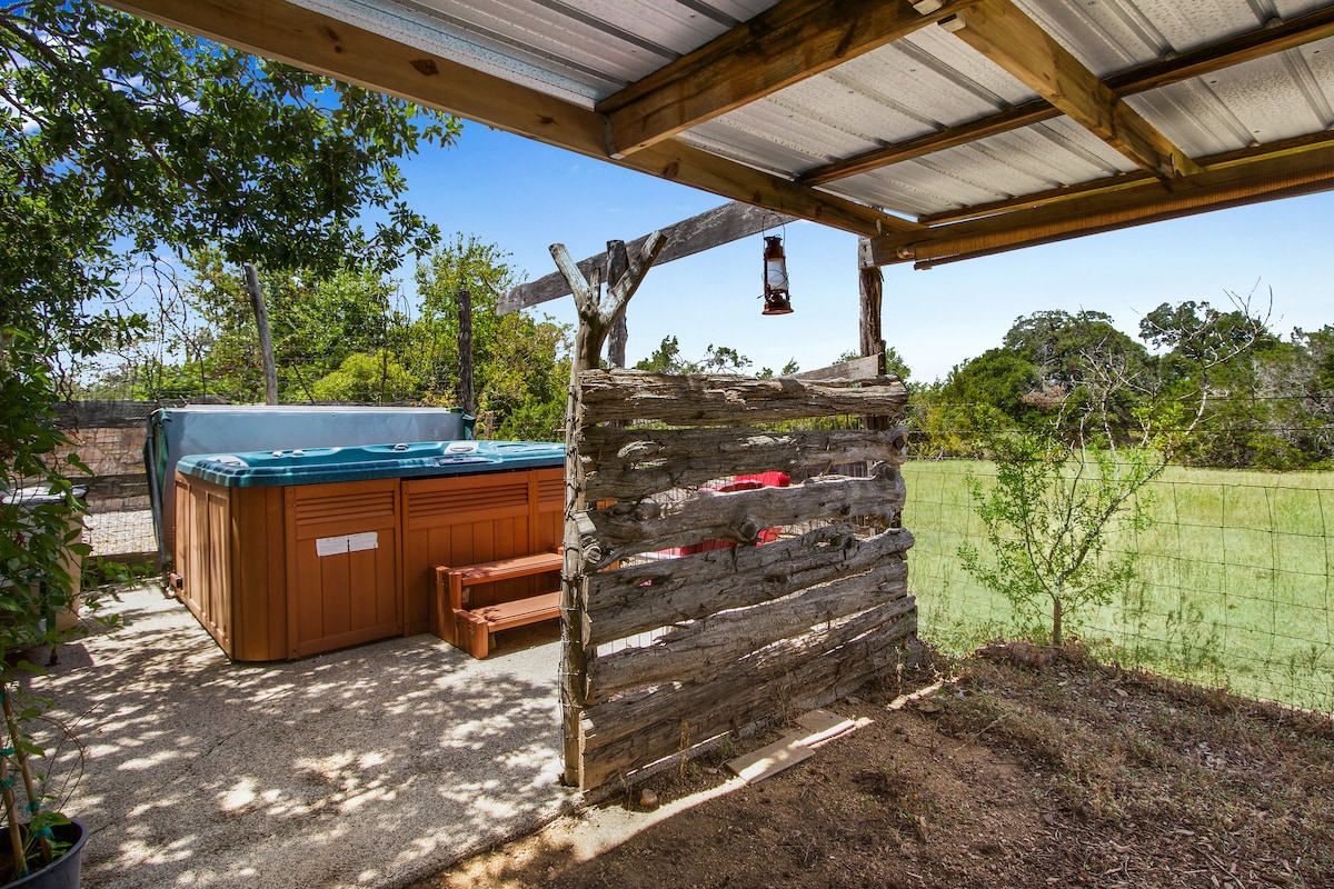 A spacious outdoor area features a hot tub nestled within a rustic wooden fence, providing a secluded space surrounded by nature. Sunlight filters through the trees, illuminating the open landscape beyond, creating a tranquil spot for relaxation.