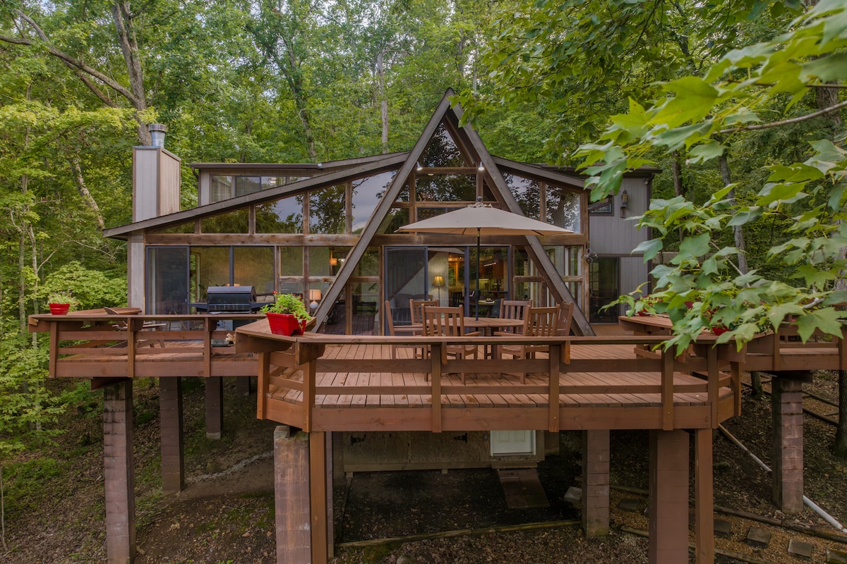 The chalet exterior features a unique architectural design with an a-frame style and large glass windows. A spacious wooden deck is visible, furnished with a dining table, umbrella, and grill, surrounded by lush greenery providing a serene atmosphere.