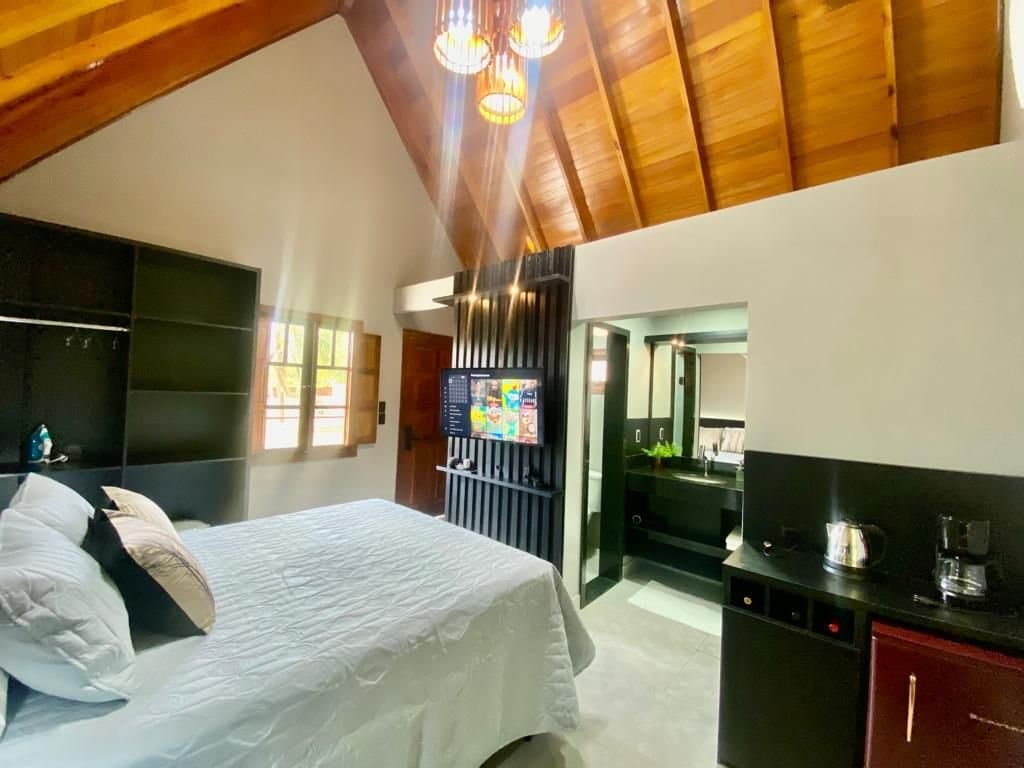 A spacious room is highlighted by a high wooden ceiling and warm lighting fixtures. A large bed with plush bedding is positioned near a wall-mounted TV, while an open doorway leads to a private bathroom, visibly equipped with modern fixtures and amenities.