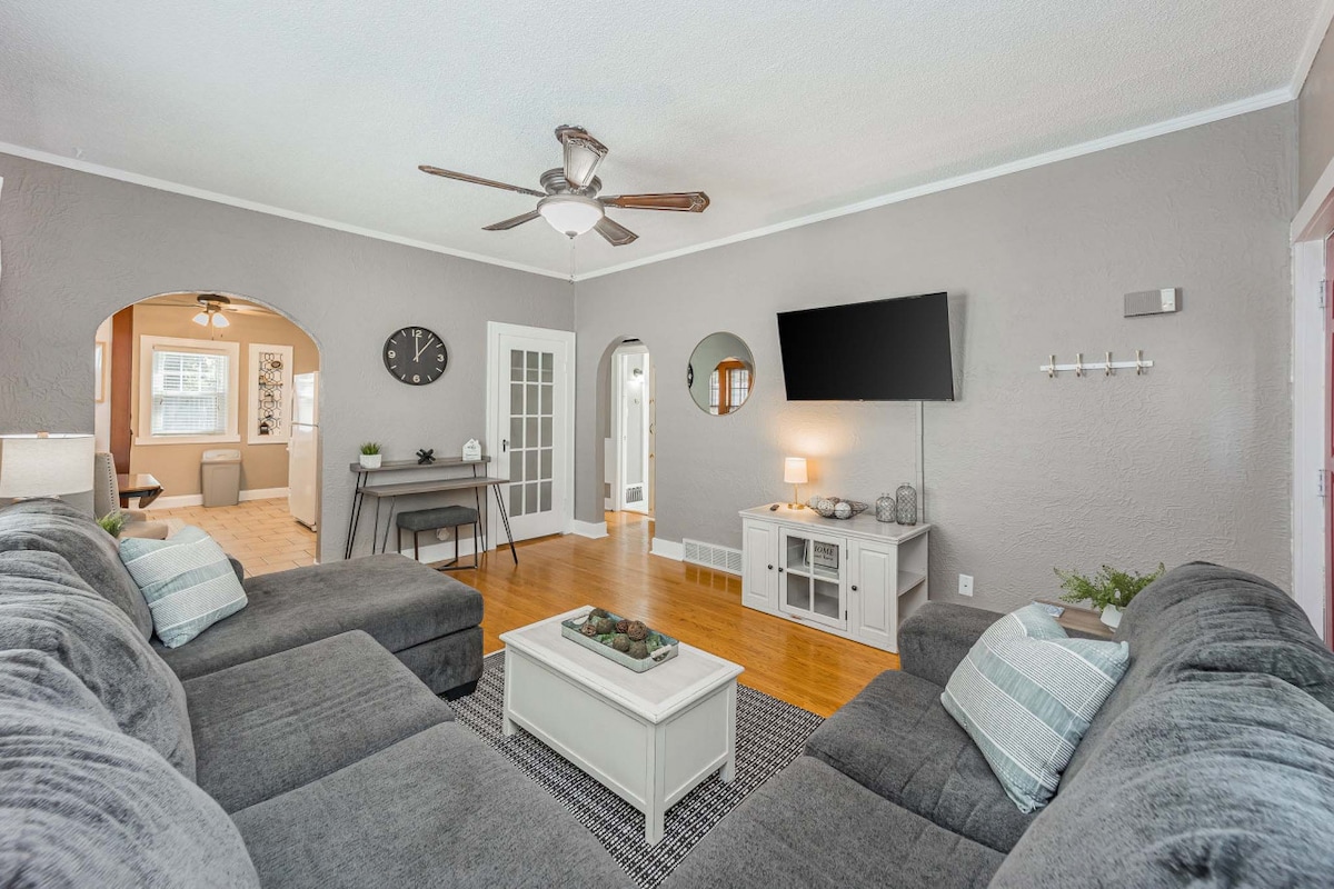 A spacious living room is showcased, featuring a plush sectional sofa with light blue and white cushions. A flat-screen television is mounted on the wall, while a coffee table positioned in front of the sofa holds decorative elements. A ceiling fan and a neutral wall color enhance the space.