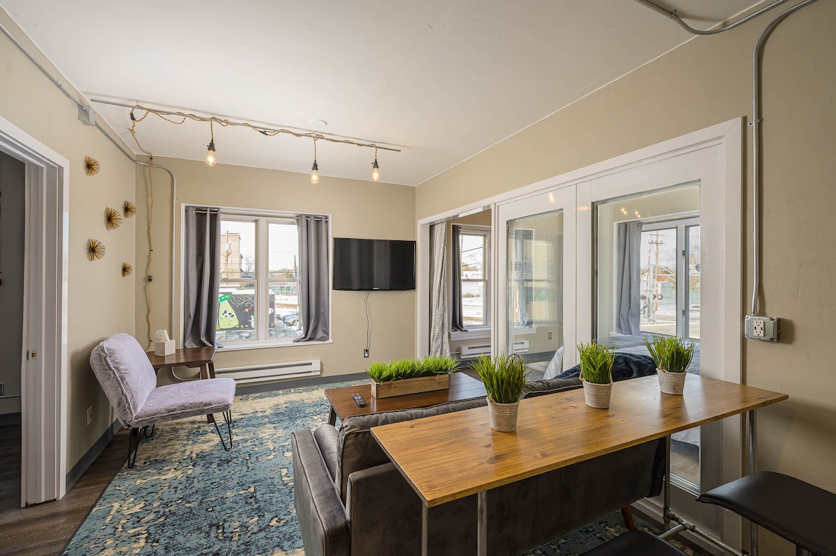 An inviting living space features large windows that allow natural light to illuminate a cozy seating area with a plush couch and a stylish chair. A wooden table is set against the wall, adorned with potted plants, while accent lighting hangs above.