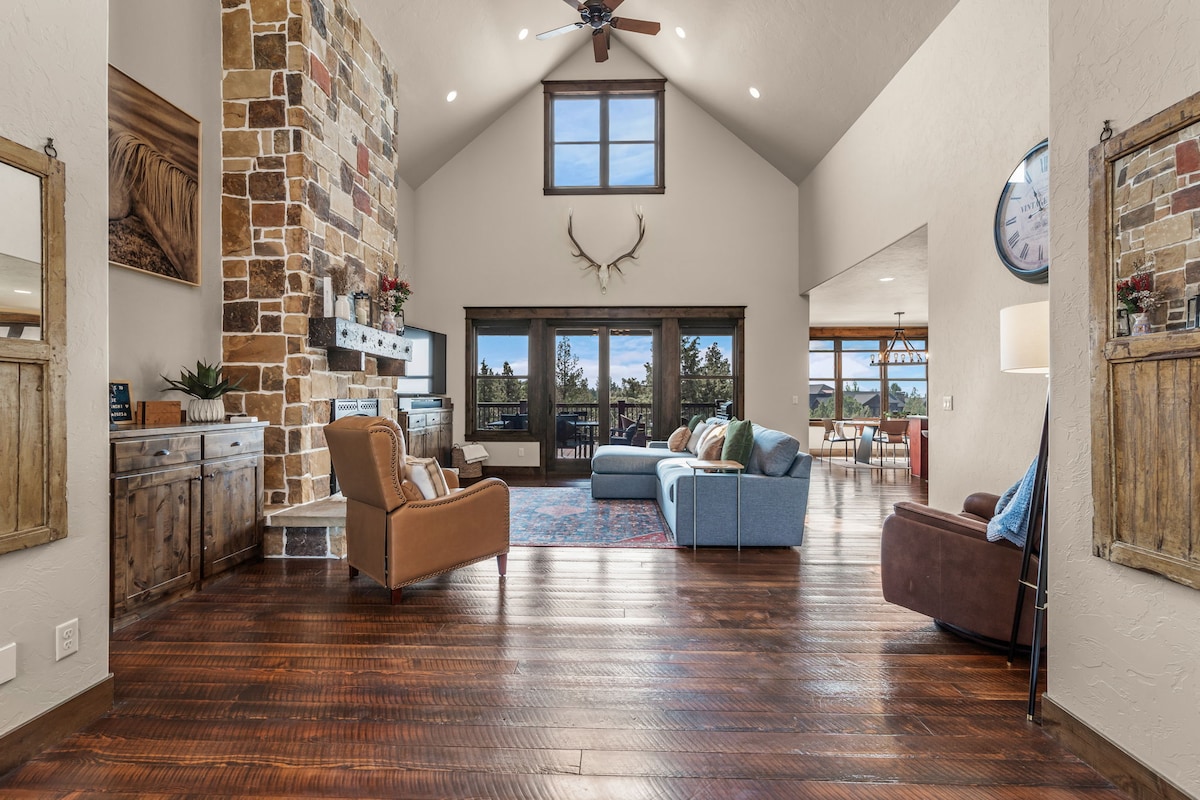 A spacious living area features a high vaulted ceiling and a stone fireplace. Large windows offer views of the surrounding landscape. Comfortable seating includes an oversized couch and armchair, arranged around a coffee table on a vibrant area rug, with natural light illuminating the wood flooring.